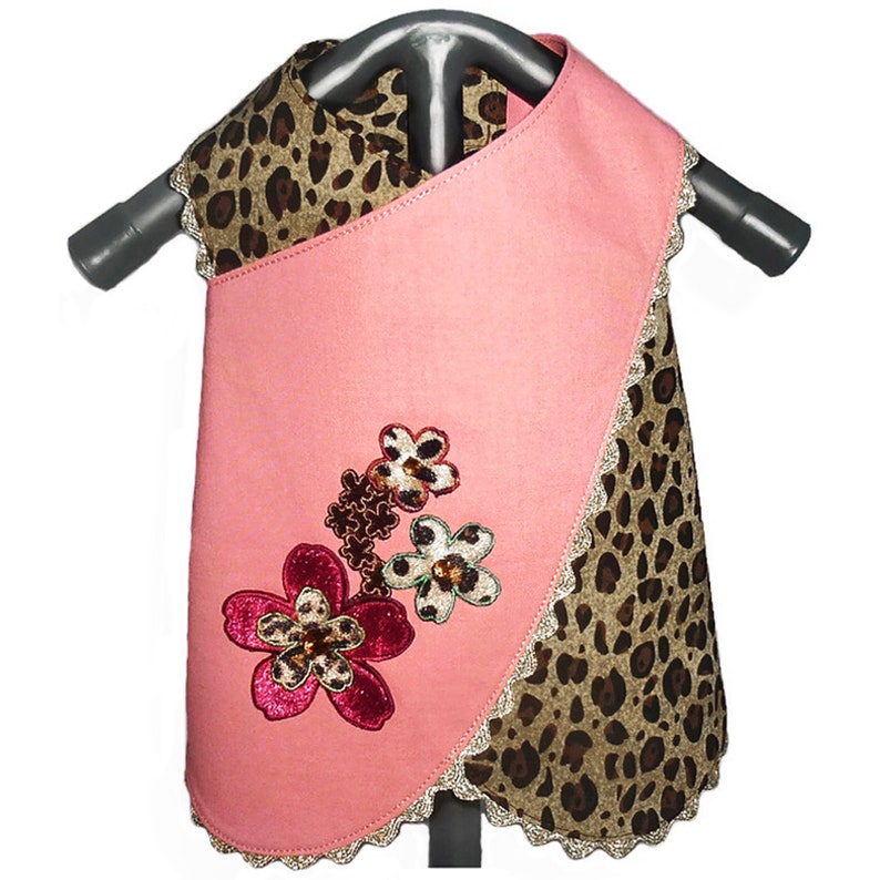 May include: A pink dog dress with a leopard print trim and a floral applique. The dress has a scalloped hem and a tie closure at the neck.