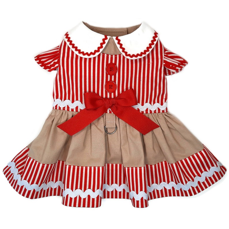 K&ouml;nnte beinhalten: Ein rot-wei&szlig; gestreiftes Hundekleid mit wei&szlig;em Kragen, roter Schleife und braunem Rock. Das Kleid hat einen Knopfverschluss und einen Ring f&uuml;r eine Leine.