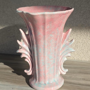 Könnte beinhalten: Eine rosa Keramikvase mit ausgestelltem Oberteil und dekorativen Seitenakzenten. Die Vase hat eine strukturierte Oberfläche mit einem dezenten blau-grünen Muster. Die Vase ist ein dekorativer Gegenstand.