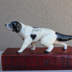 Puede incluir: Una figurita de cerámica blanca y negra de un perro de caza en posición de carrera. La figurita está de pie sobre un libro rojo con letras doradas. La figurita tiene una etiqueta en su lado que dice "Bilboy 204".