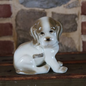 May include: A white and brown ceramic figurine of a sitting dog. The dog has a brown nose and brown ears. The figurine has a label on the bottom that reads "Berolina, Germany".