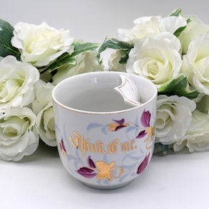 May include: A white porcelain cup with a gold rim and floral design. The cup has a unique handle that is shaped like a small spout. The cup is decorated with purple and gold flowers and the words "Think of me." in gold lettering.