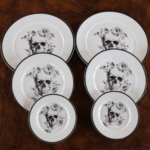 May include: A set of white ceramic plates with a black rim. Each plate features a black and white skull design surrounded by floral elements. The set includes plates of varying sizes, suitable for a dining table setting.