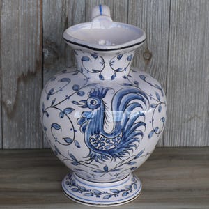 May include: A white ceramic pitcher with a blue rooster design and leaf accents. The pitcher has a rounded body, a flared spout, and a small handle. The base is a pedestal shape. The pitcher is approximately 10 inches tall.