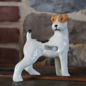 Könnte beinhalten: Keramikhundefigur mit weißem Körper, schwarzem Fleck und hellbraunen Ohren. Die Figur hat ein kleines, rechteckiges Etikett mit der Aufschrift "Bowser -109". Der Hund steht auf einer Holzoberfläche.