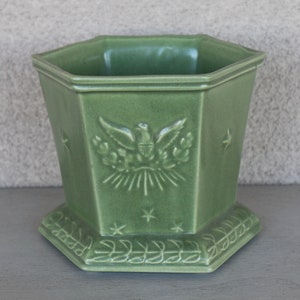 Può includere: Un vaso da fiori esagonale in ceramica verde con un designo in rilievo di un'aquila con le ali spiegate, uno stendardo con stelle e un raggio di sole. Il vaso ha un bordo decorativo attorno alla parte superiore e inferiore.