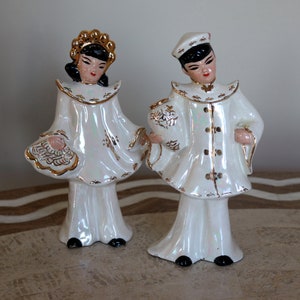 Puede incluir: Dos figuritas de porcelana de un hombre y una mujer con ropa tradicional china. La mujer lleva un vestido blanco con detalles dorados y una corona dorada. El hombre lleva una túnica blanca con detalles dorados y un sombrero negro. Ambas figuritas tienen el pelo negro y rasgos faciales pintados.