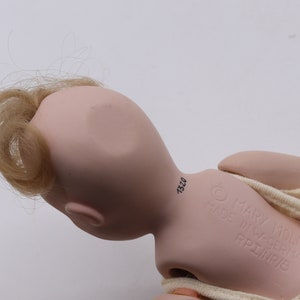 May include: A vintage doll with a pale pink complexion, blonde hair, and a cream-colored outfit. The doll's head is turned, revealing the back of its head and the inscription "MARY MOLINE MADE IN W-GERMANY RPI/NR/6".