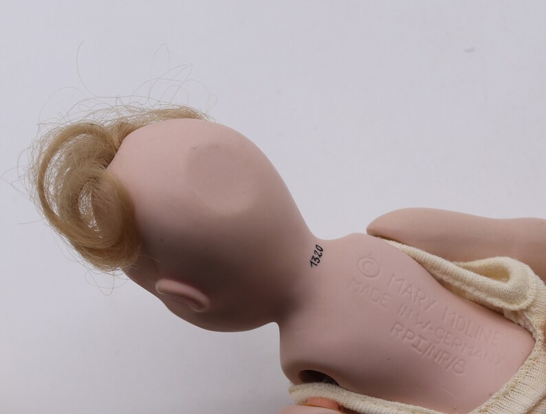 May include: A vintage doll with a pale pink complexion, blonde hair, and a cream-colored outfit. The doll's head is turned, revealing the back of its head and the inscription "MARY MOLINE MADE IN W-GERMANY RPI/NR/6".