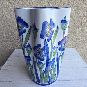 Può includere: Un vaso in ceramica bianca con bordo ondulato e smerlato, caratterizzato da un motivo floreale dipinto a mano di iris blu con steli e foglie verdi. Il vaso ha una forma cilindrica affusolata e una finitura liscia e lucida con bordo blu.