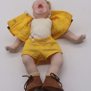 May include: A vintage porcelain doll with blonde hair, wearing a yellow jacket and shorts, a white ribbed undershirt, and brown leather shoes. The doll's mouth is open, and it appears to be crying. The doll is lying on a white surface.