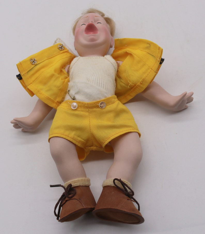 May include: A vintage porcelain doll with blonde hair, wearing a yellow jacket and shorts, a white ribbed undershirt, and brown leather shoes. The doll's mouth is open, and it appears to be crying. The doll is lying on a white surface.