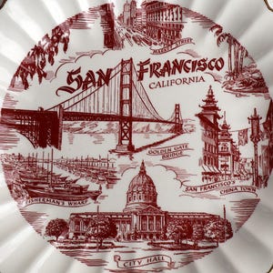 Könnte beinhalten: Ein roter und weißer Keramikteller mit einem Design von Sehenswürdigkeiten in San Francisco, darunter die Golden Gate Bridge, Fisherman's Wharf, das Rathaus und Chinatown. Der Teller hat einen gewellten Rand und der Text "San Francisco, Kalifornien" ist in Rot gedruckt.