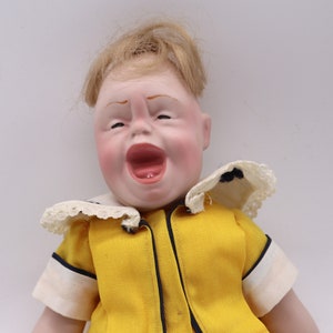 May include: A vintage doll with a detailed, expressive face, open mouth, and blonde hair. The doll wears a yellow outfit with a white collar and black trim. The doll's face is painted with rosy cheeks and red lips.