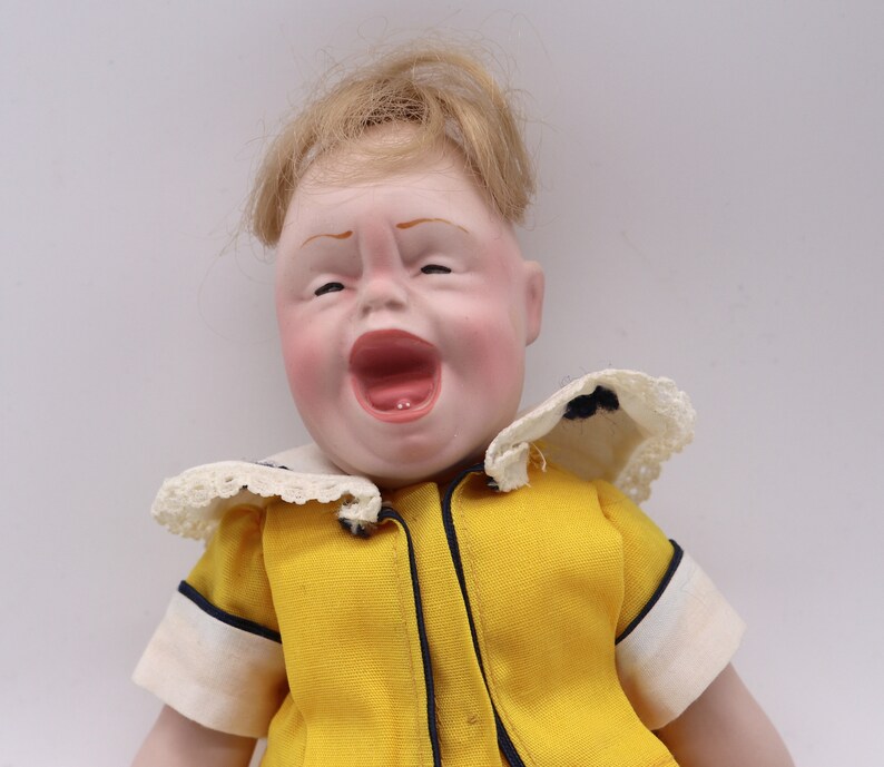 May include: A vintage doll with a detailed, expressive face, open mouth, and blonde hair. The doll wears a yellow outfit with a white collar and black trim. The doll's face is painted with rosy cheeks and red lips.