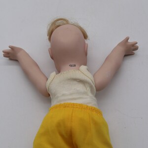 May include: A porcelain doll with blonde hair and outstretched arms, wearing a cream-colored tank top and yellow shorts. The doll's neck has the number "1320" printed on it.