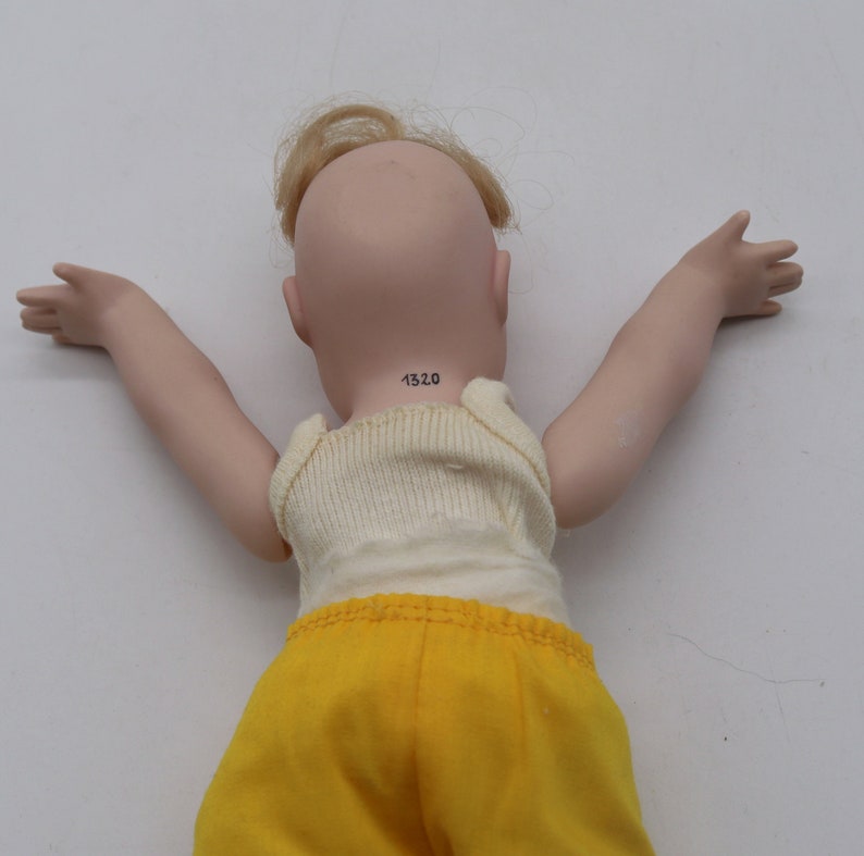 May include: A porcelain doll with blonde hair and outstretched arms, wearing a cream-colored tank top and yellow shorts. The doll's neck has the number "1320" printed on it.