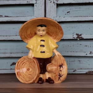 Puede incluir: Una figurita de cerámica de una persona que lleva una camisa amarilla y un sombrero cónico marrón, sosteniendo dos cestas marrones. La figurita está de pie sobre una superficie de madera.