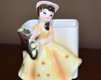 May include: A vintage ceramic planter shaped like a girl in a yellow dress with white polka dots. She is holding a brown basket with green flowers. The planter is white with a square base.