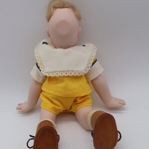 May include: A vintage doll with blonde hair, wearing a yellow shorts set and brown shoes. The doll has a cream-colored collar with lace trim and navy blue accents. The doll is sitting with its arms and legs extended.