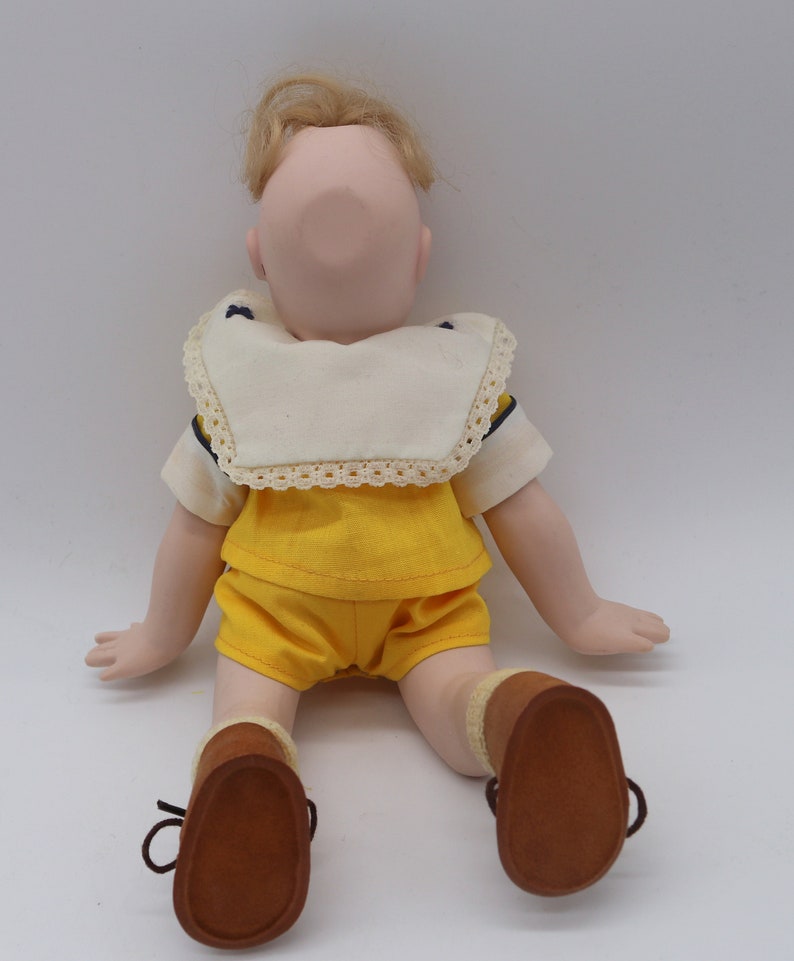 May include: A vintage doll with blonde hair, wearing a yellow shorts set and brown shoes. The doll has a cream-colored collar with lace trim and navy blue accents. The doll is sitting with its arms and legs extended.
