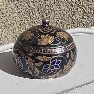 May include: A small, round, black metal lidded container featuring intricate hand-painted floral patterns in gold, blue, and white enamel. The lid has a central knob and a geometric grid design at the top, with a silver-toned metal rim.