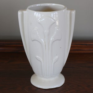 May include: White ceramic vase with a stylized floral design. The vase has two handles and a rounded base.