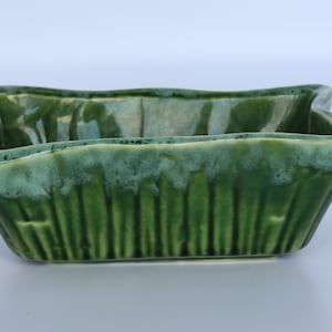 May include: A rectangular, ceramic planter with a unique, star-shaped rim. The planter is glazed in shades of green, with a darker green base and a lighter, turquoise-green rim. Vertical ridges add texture to the sides.
