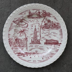 May include: White ceramic plate with a red illustration of Daytona Beach, Florida landmarks, including the Clock Tower and the Ocean Pier. The plate features a decorative rope-like border and text that reads "Worlds Most Famous Beach."