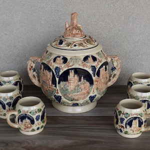 Puede incluir: Un tazón grande de ponche de cerámica con un diseño de castillo y vides. El tazón está rodeado de siete tazas de cerámica más pequeñas con diseños similares. Las tazas y el tazón son todos en tonos beige, marrón, azul y verde.