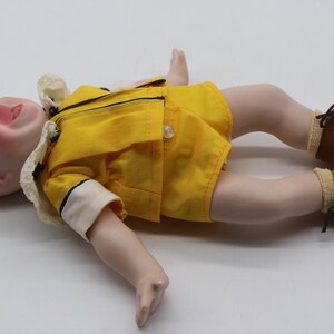 May include: A vintage doll with a porcelain face, blonde hair, and rosy cheeks. The doll wears a yellow outfit with black trim, white collar, and brown shoes. The doll is lying on its back with its mouth open.