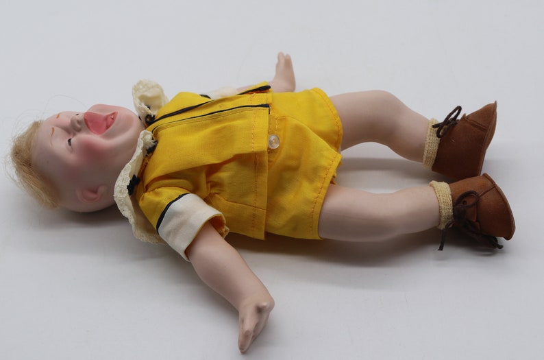 May include: A vintage doll with a porcelain face, blonde hair, and rosy cheeks. The doll wears a yellow outfit with black trim, white collar, and brown shoes. The doll is lying on its back with its mouth open.