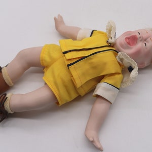 May include: A vintage doll with a porcelain face and blonde hair, dressed in a yellow outfit with black trim. The doll is wearing brown shoes and is lying on its back with its mouth open.