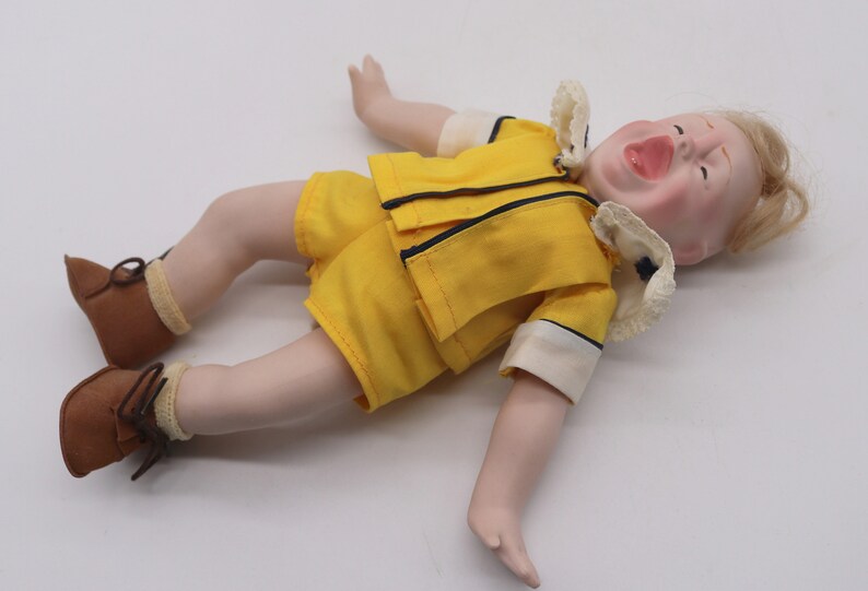 May include: A vintage doll with a porcelain face and blonde hair, dressed in a yellow outfit with black trim. The doll is wearing brown shoes and is lying on its back with its mouth open.