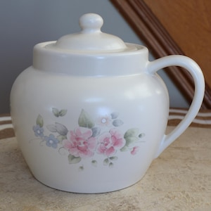 Op de afbeelding: Witte keramische pot met een deksel en een handvat. De pot heeft een bloemenmotief met roze, blauwe en groene bloemen.