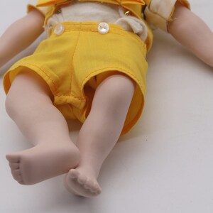 May include: A porcelain doll with a cream-colored top, yellow shorts, and matching suspenders. The doll's arms and legs are extended, with detailed hands and feet. The doll's outfit is accented with white buttons.
