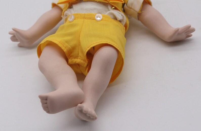 May include: A porcelain doll with a cream-colored top, yellow shorts, and matching suspenders. The doll's arms and legs are extended, with detailed hands and feet. The doll's outfit is accented with white buttons.