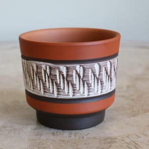 May include: A terracotta planter with a brown and white geometric pattern. The planter has a wide base and a narrow top.