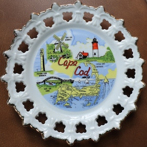 Può includere: Un piatto decorativo bianco con bordo dorato e una mappa dettagliata di Cape Cod, Massachusetts. Il piatto presenta illustrazioni di un mulino a vento, un faro e un ponte. Le parole "Cape Cod" sono ben visibili.