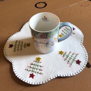 Może przedstawiać: Ceramiczny kubek z zimową sceną i jasnoniebieskim uchwytem stoi na białej filcowej podkładce. Podkładka posiada haftowane wzory choinek z czerwonymi i ż&oacute;łtymi akcentami gwiazd i falistą krawędzią.