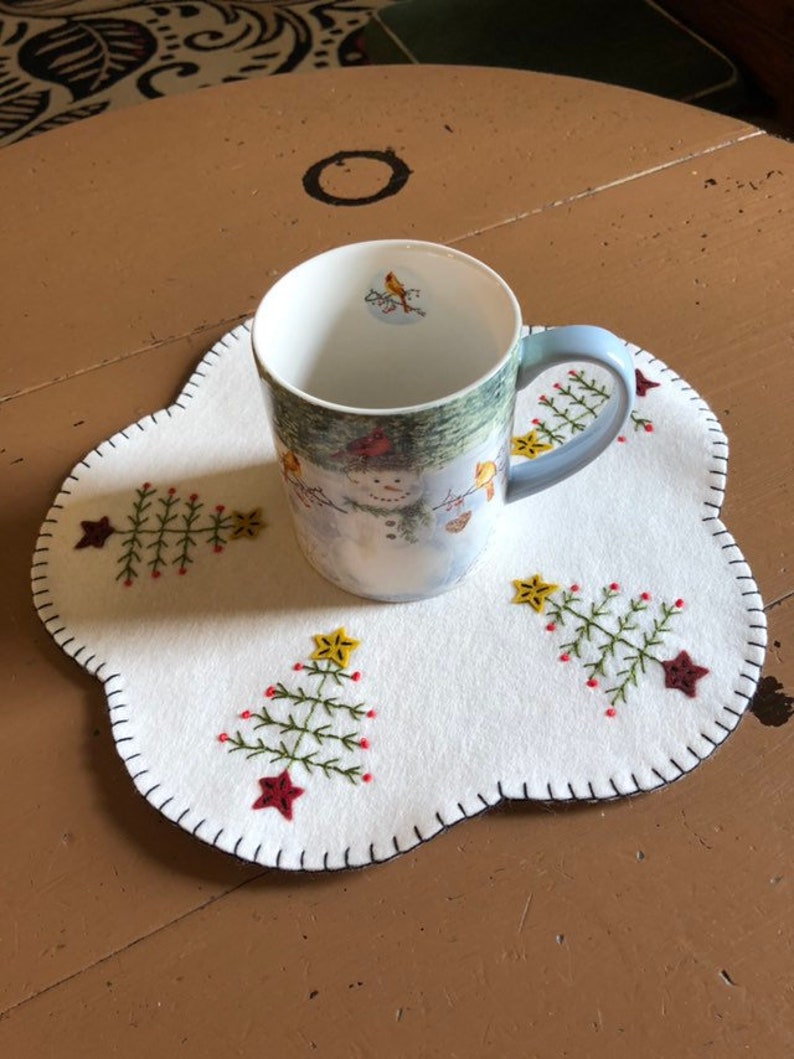 Może przedstawiać: Ceramiczny kubek z zimową sceną i jasnoniebieskim uchwytem stoi na białej filcowej podkładce. Podkładka posiada haftowane wzory choinek z czerwonymi i ż&oacute;łtymi akcentami gwiazd i falistą krawędzią.