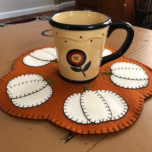 Wool felt primitive pumpkin candle mat with an orange background measuring 12 inches across with off white pumpkins in the scallops.