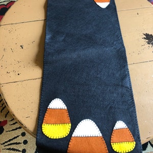 May include: Black felt table runner with five felt candy corn appliques. The candy corn appliques are yellow, orange, and white.