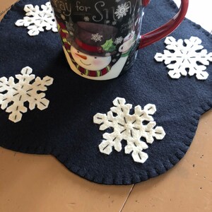 May include: A black felt snowflake-shaped coaster with white felt snowflakes on it. A white mug with a snowman design and the text "Pray for Snow" is sitting on the coaster.