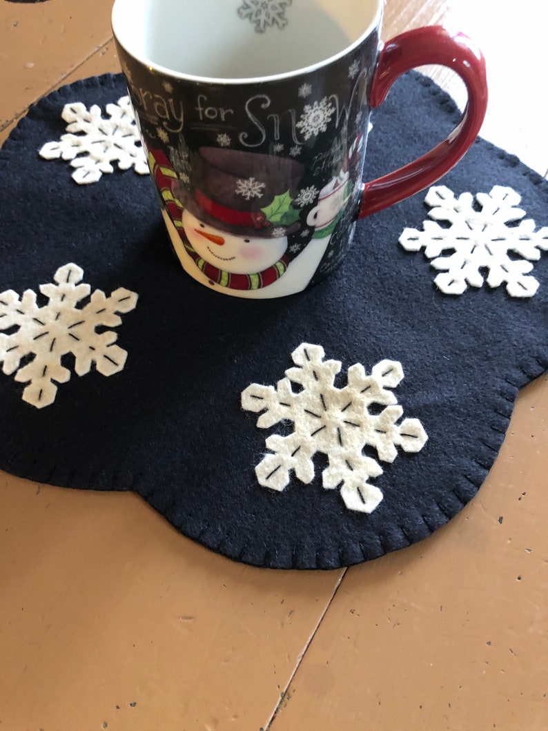 May include: A black felt snowflake-shaped coaster with white felt snowflakes on it. A white mug with a snowman design and the text "Pray for Snow" is sitting on the coaster.