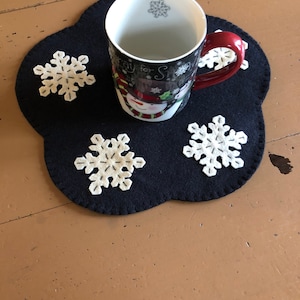 May include: A black felt snowflake-shaped coaster with four white felt snowflakes. A white mug with a snowman design and the text "Ready for Snow" is on top of the coaster.