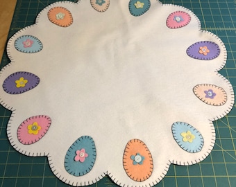 Easter egg table mat measuring 22 inches across with an off white background with pastel eggs in the scallops.