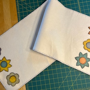 May include: A white felt table runner with a black stitched border and colorful felt flower appliques. The runner is folded in half and laid on a green cutting mat.