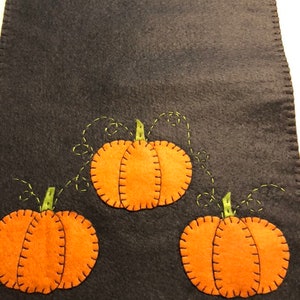 May include: A black felt table runner with three orange felt pumpkins with green felt stems and vines. The pumpkins have a stitched outline and a stitched line down the center.
