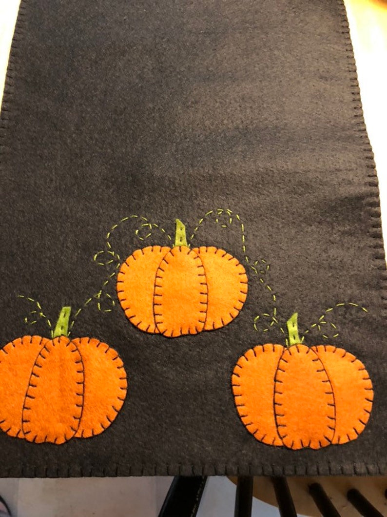 May include: A black felt table runner with three orange felt pumpkins with green felt stems and vines. The pumpkins have a stitched outline and a stitched line down the center.